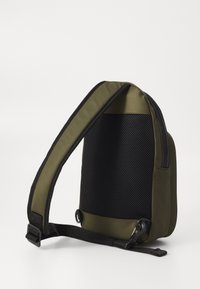 Green and black crossbody bag featuring a smooth fabric, a mesh panel, a padded strap, and metal hardware. Compact design with an ergonomic shape.