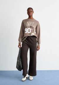 Brūna Nike džemperis ar baltām grafikām, apģērbts ar tumšiem, brīvākas formas biksēm un baltām sporta apaviem. Modelis tur teksturētu pelēku somu.