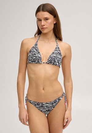 Jonge vrouw met een zwart-witte zebraprint halterbikini met roze accenten, staand tegen een effen lichte achtergrond.