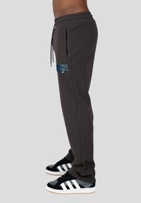 Graue Sweatpants mit einem Kordelzugbund und Seitentaschen, mit "WRONG FRIENDS" in Blau auf der Seite gestickt, getragen mit schwarzen Sneakers.