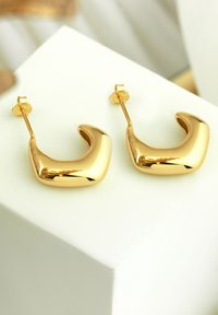 Guld-tonade örhängen har en slät, böjd design med en polerad yta. De har en öronkrok och en chunky, abstrakt form.