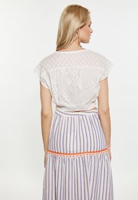 Fehér, texturált crop top, kapucnis ujjal; hosszú, csíkozott szoknyával párosítva, amely lila és narancssárga színű kiegészítőkkel rendelkezik.