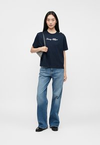 Camiseta de algodón azul marino con el texto blanco "Tommy Hilfiger", combinada con unos jeans acampanados de color azul claro y un bolso pequeño de diseño estampado.