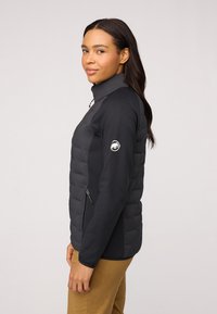 Svart vadderad jacka med en mjuk textur, hög krage, dragkedjefickor på sidan och en vit logotyp på vänster ärm.