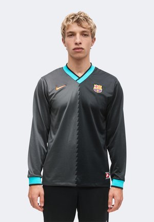Joven con cabello rubio corto que lleva una camiseta de fútbol de manga larga del FC Barcelona en negro y gris con cuello y puños turquesa.