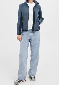 Veste zippée bleu marine avec motif texturé, poches latérales et jean bleu clair. Le mannequin porte des baskets grises avec des accents rouges.
