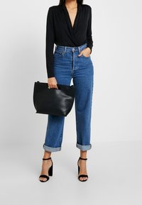 Schwarzes langärmeliges Wickeloberteil, hochtaillierte blaue Jeans mit umgeschlagenen Säumen und schwarze Ledertasche mit Griffen. Schwarze Riemchen-High Heels.