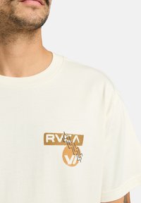Cream-colored cotton t-shirt with a round neckline. Features a screen print logo in orange and black on the chest. Casual design.