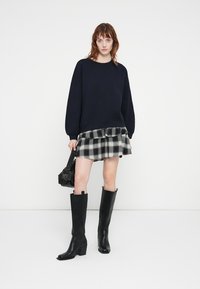 Pull oversized marine, superposé sur une mini-jupe à carreaux noirs et blancs avec un ourlet volanté, associé à des bottes noires à talons hauts jusqu'aux genoux.