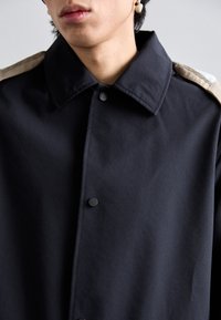 Primer plano de una persona que lleva un abrigo negro abotonado con acentos beige en los hombros y un pendiente beige en la oreja izquierda.