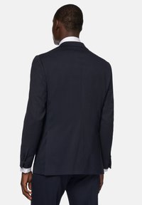 Marinblå kavaj med texturerad yta; enkel sprund bak, två knappar på varje ärm, och en vit skjorta synlig under.