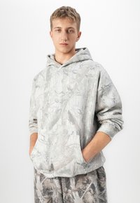 Giovane uomo che indossa un hoodie mimetico grigio e beige e pantaloni abbinati, con le mani nella tasca frontale, in piedi su uno sfondo chiaro.