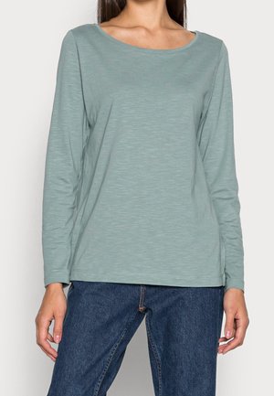 Kvinde iført en lysegrøn langærmet rundhalset trøje og blå denimjeans, stående med hænderne afslappet ned langs siderne.