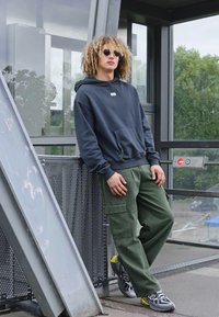 Hoodie azul-marinho com bolso frontal, calças cargo verdes e óculos de sol pretos. De pé numa plataforma com corrimãos de metal e árvores nas proximidades.