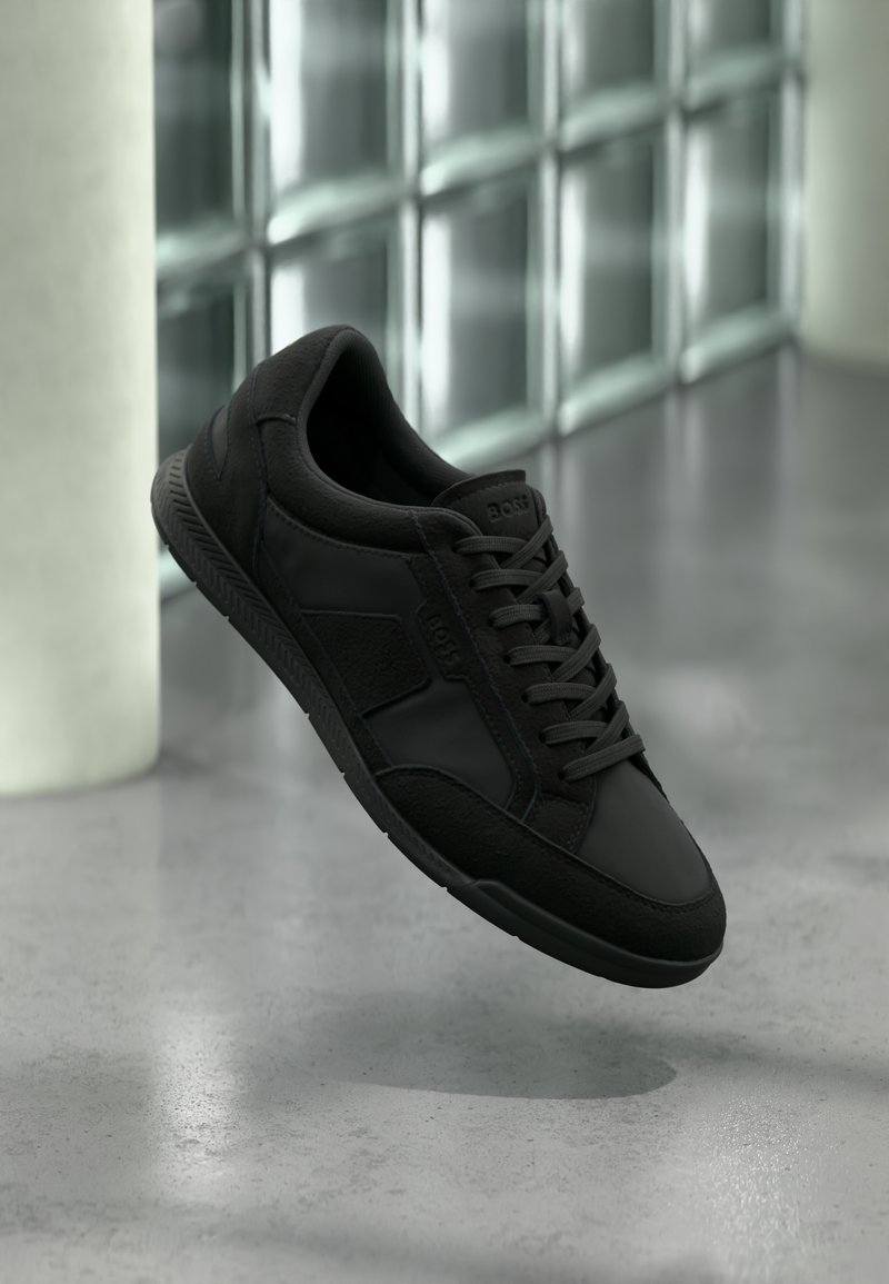 Baskets noires BOSS avec panneaux texturés et lacets, suspendues au-dessus d'un sol en béton poli près de blocs de verre et d'une colonne blanche.