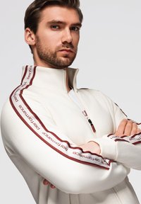 Chaqueta blanca con cremallera y cuello alzado, con acentos en color burdeos y texto a lo largo de las mangas, fabricada con un tejido suave y de diseño ajustado.