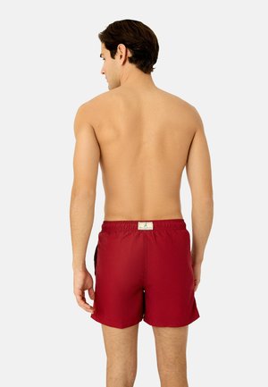 Mann mit kurzen dunklen Haaren, oberkörperfrei, trägt rote Badeshorts, steht mit dem Rücken zur Kamera vor einem schlichten, hellen Hintergrund.