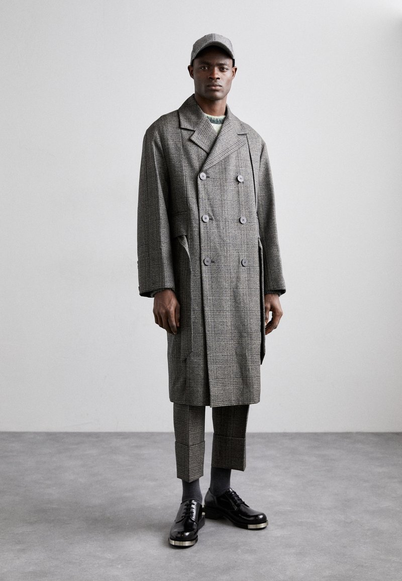 Cappotto doppiopetto grigio a quadri con grandi bottoni, abbinato a pantaloni a quadri coordinati e scarpe nere con lacci. Un berretto grigio completa il look.