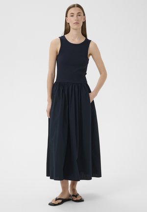 Žena stojaca s jednou rukou v kapsi, oblečená v čiernych bezrukávoch maxi šatách a čiernych sandáloch na bielom pozadí.