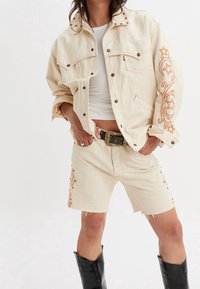 Béžová džínová bunda s oversized střihem a bronzovými cvoky na límci, doplněná o odpovídající šortky s vyšívanými detaily a třepenými lemy.