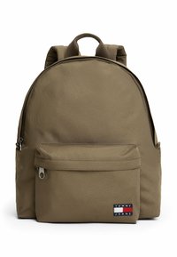 Mochila sintética marrón con forma redondeada, correas de hombro acolchadas, un bolsillo frontal con cremallera y un parche con el logo en la parte inferior delantera.