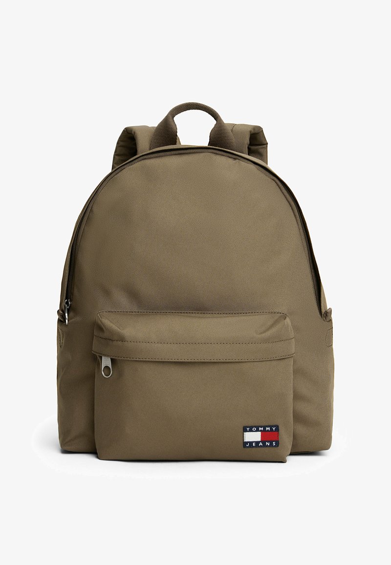 Zaino sintetico marrone con forma arrotondata, spallacci imbottiti, tasca frontale con zip e una toppa con logo sulla parte anteriore inferiore.