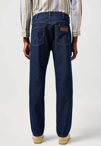 Jeans de mezclilla en azul oscuro, con un corte relajado, costuras visibles y dos bolsillos traseros con un patrón decorativo y una etiqueta de logo.