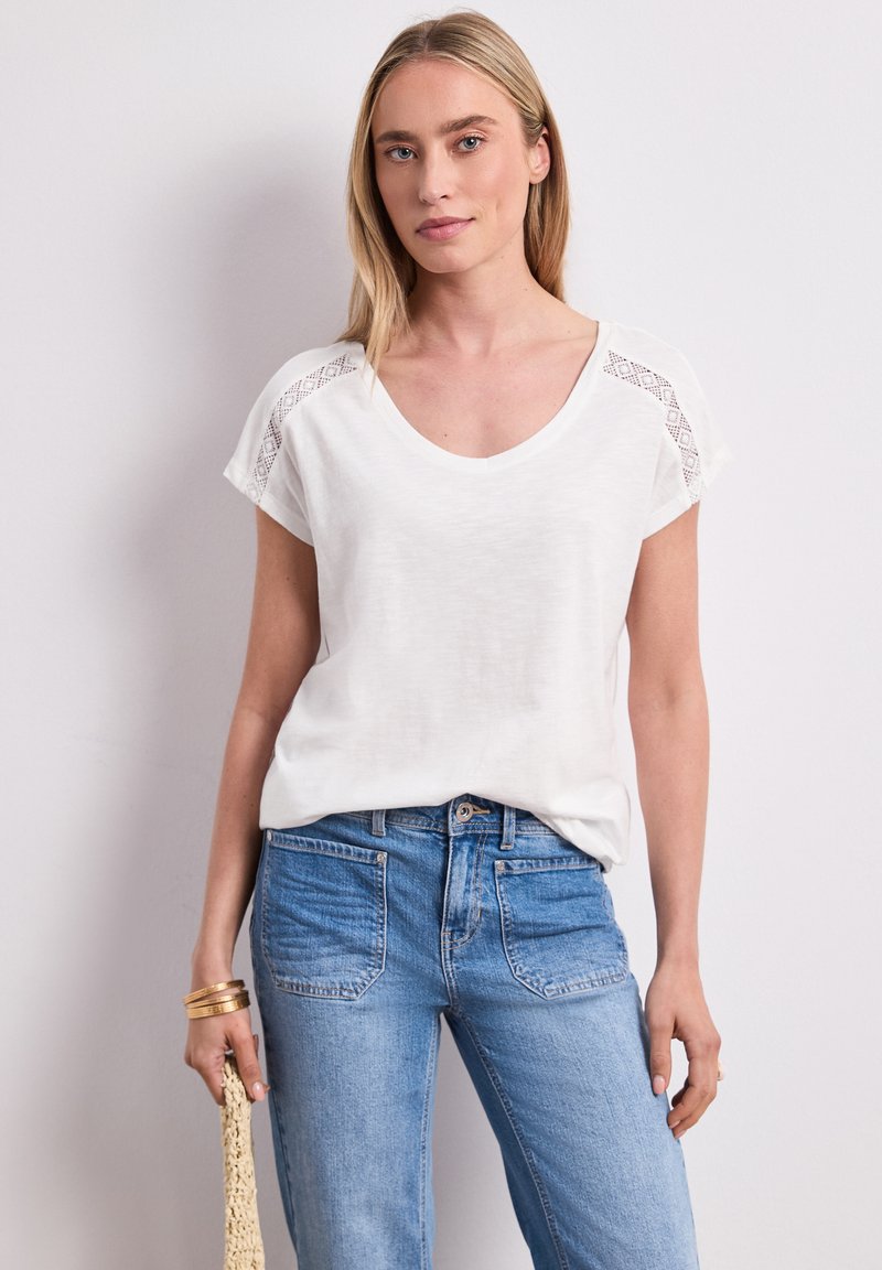 Femme aux cheveux blonds porte un top blanc à manches courtes avec un détail en dentelle sur les épaules, un jean bleu, des bracelets dorés, et tient un sac tissé beige.