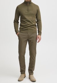 Muž v olivově zeleném svetru s kratkým zipem, odpovídajících olivových kalhotách a béžových semišových kotníkových botách, stojí s jednou rukou v kapse.