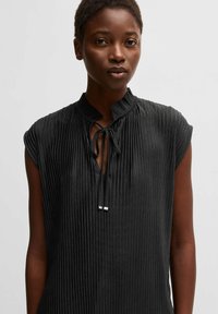 Blusa nera senza maniche in tessuto testurizzato e rigato. Presenta un colletto alla mandarina e un dettaglio con fiocco al collo con inserti metallici.