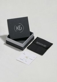 Svart presentförpackning med silver- och vit logotyp, som innehåller en mindre tygpåse och etiket, med text och en guldaccent.