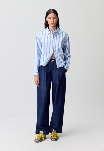 Camisa a rayas azules, corte holgado, con cuello abotonado; pantalones de mezclilla oscuros y anchos; zapatillas metálicas con cordones y detalles amarillos; hebilla de cinturón dorada.