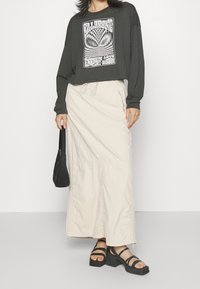 Donna che indossa una maglietta nera a maniche lunghe con grafica, una maxi gonna beige chiaro, sandali platform neri e porta una borsa nera.