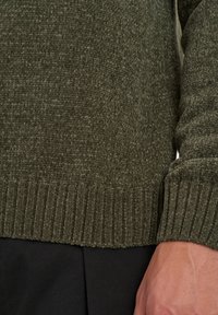 Maglione a maglia verde oliva, orlo e polsini a coste, tessuto testurizzato, vista ravvicinata che mostra i dettagli. Indossato sopra un paio di pantaloni in tessuto neri.