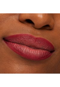 Dyp rød matt leppestift på fyldige lepper, med synlig tekstur og en gradienteffekt mot kantene, mot en jevn brun hudbakgrunn.