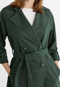 Dwurzędowy zielony płaszcz trench z dużymi klapami, wiązanym w pasie i bocznymi kieszeniami; wykonany z gładkiego materiału o matowym wykończeniu.