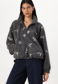 Casaco de fleece cinza com gola alta, com bordados florais e fecho na frente. O tecido tem um aspeto suave e texturizado.