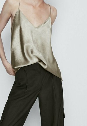 Kobieta prezentująca złoty satynowy top na cienkich ramiączkach, wsunięty w czarne, szerokie spodnie z wysokim stanem na jednolitym tle.