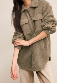 Olijfgroene, donzige fleecejas met corduroy accenten en twee voorzakken. Heeft een knoopsluiting en opgerolde mouwen.