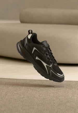 Zapatilla deportiva negra y gris con detalles de malla y sintéticos, cordones atados, flotando sobre una alfombra beige en un ambiente interior con un sofá de fondo.