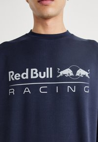 Tamsiai mėlynas apvalaus kaklo džemperis su ryškiu baltu "Red Bull Racing" logotipu ir dviem bulių grafikomis priekyje.