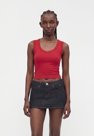 Kvinde med fletninger iført en rød ærmeløs crop top og en sort denim mininederdel, stående mod en ensfarvet hvid baggrund.