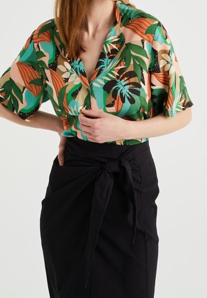 Kobieta nosząca bluzkę z krótkim rękawem i tropikalnym liściastym wzorem, wsadzoną w czarną spódnicę z wysokim stanem z detalem węzła z przodu.