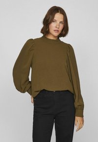 Olivgrön långärmad blus med puffärmar och hög halsringning, kombinerad med svarta jeans. Texturerad tyg, avslappnad passform.