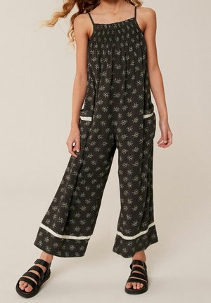 Combinaison noire à motifs avec fines bretelles, jambes larges et amples, bordure blanche sur les poches, et détails floraux sur l'ensemble. Associée à des sandales noires.