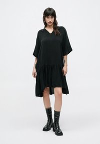 Μαύρο φόρεμα μήκους μέχρι το γόνατο με φαρδιά ρεβέρ, V-λαιμόκοψη και κοντά μανίκια. Μαλακή υφή, μινιμαλιστικός σχεδιασμός, σετάρεται με μαύρες δερμάτινες μπότες.