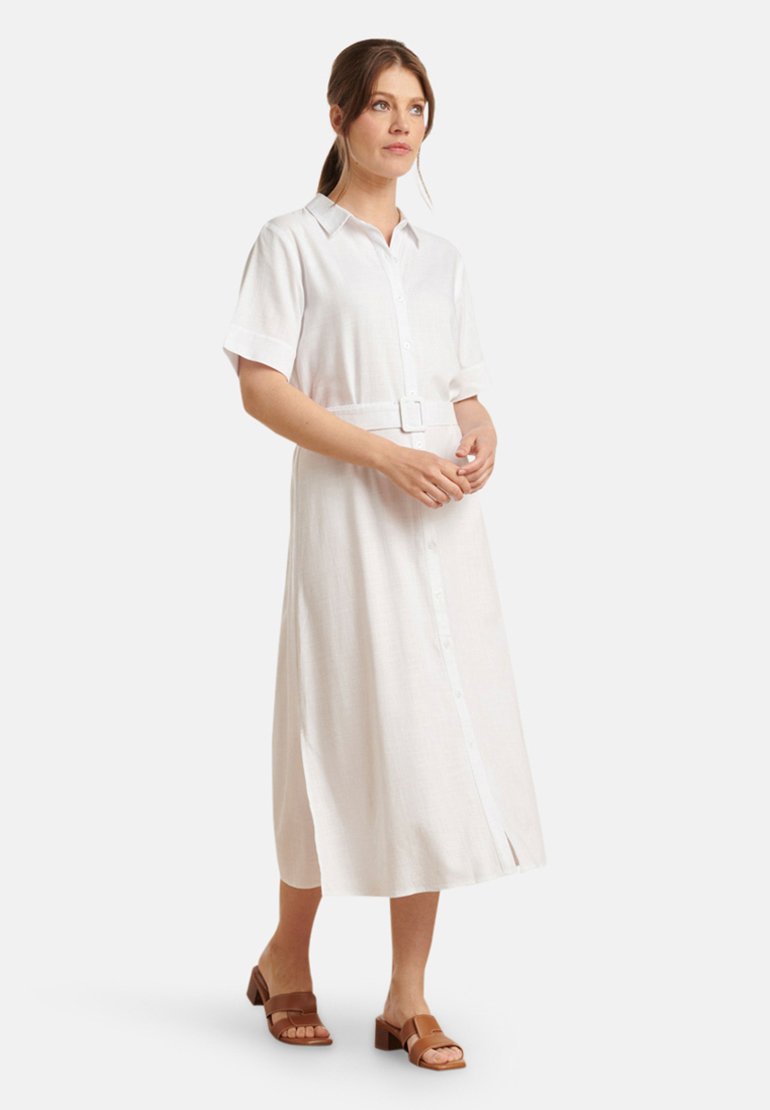 Witte linnen jurk met korte opgerolde mouwen, knopen voorkant, ceintuur in de taille en zijsplit, gedragen met bruine slide sandalen.