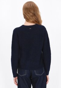Mørkeblå striktrøje med en blød tekstur, rund halsudskæring og lange ærmer. Har et lille logo på ryggen ved halsudskæringen. Sammen med blå denim.