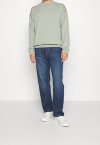 Lichtgroene trui met ronde hals en ribbels aan de mouwen, gecombineerd met blauwe jeans. Model draagt witte sneakers en staat voor een neutrale achtergrond.
