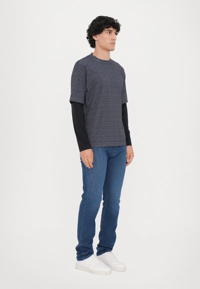 Tumšzilgs īsām piedurknēm tops ar teksturētu ģeometrisku rakstu, virsū uzvilkts uz garām piedurknēm melna krekla, apvienots ar zilām džinsām un baltām sporta apaviem.